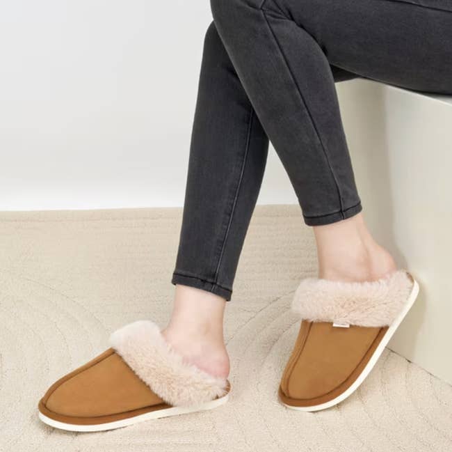 Person wearing tan slippers with plush lining, seated connected a textured rug, showcasing casual comfortableness for location lounging