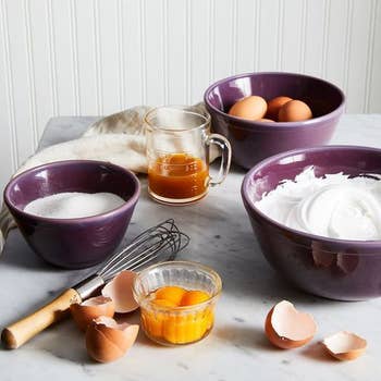 Baking ingredients on a counter, including eggs, flour, sugar, and honey, with mixing bowls and a whisk