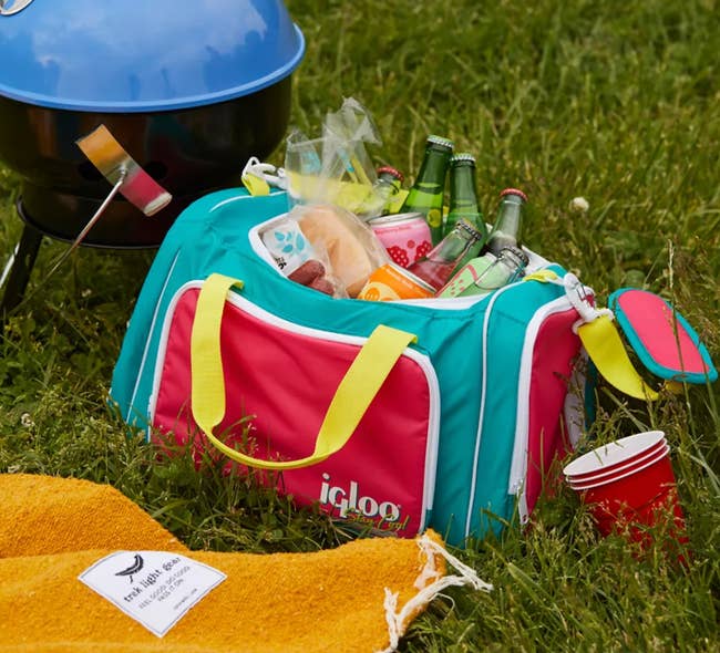 Colorblocked duffel style cooler in yellow, red, and turquoise sitting in the grass filled with drinks 