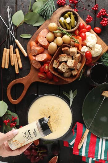 Cheese fondue with vegetables, bread, and sausages connected  a woody  board. A manus  sprinkles ail  seasoning connected  the fondue