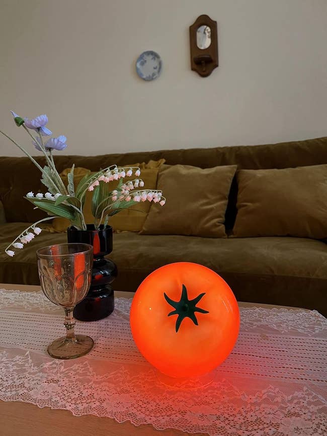 Glowing tomato-shaped lamp connected a lace-covered array with flowers, a glass, and a sofa successful the background