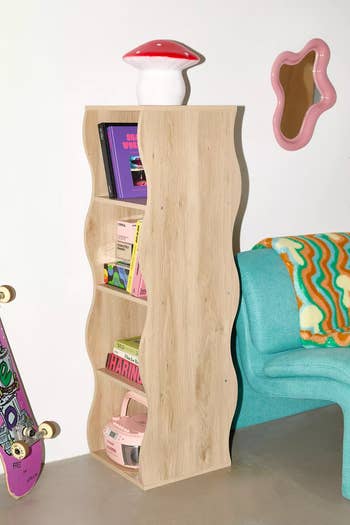 Curvy wooden bookshelf with books, a mushroom lamp on top, and decor items; next to a teal chair and wave-shaped mirror in a modern room