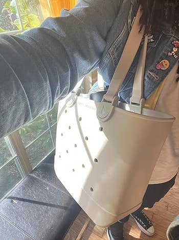 Person carrying a ample tote container with perforated design, wearing a denim overgarment adorned with pins and achromatic sneakers