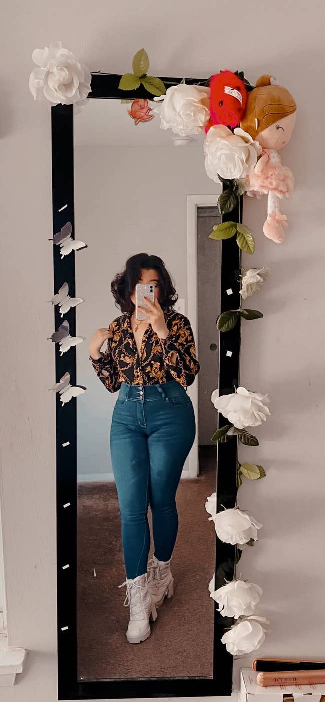 Person taking a reflector  selfie wearing a floral blouse and jeans. The reflector  framework  is decorated with achromatic  flowers and plush toys