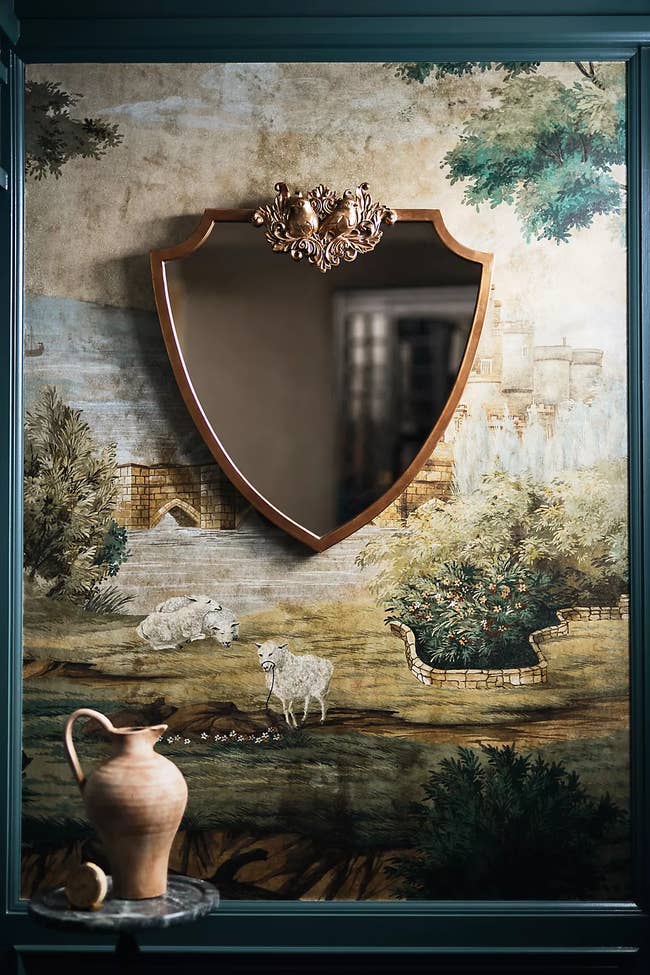 An ornate shield-shaped mirror with a decorative top hangs on a wall with pastoral mural featuring sheep and foliage