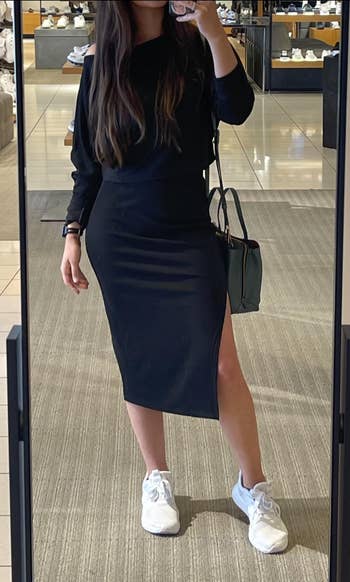 Person in a store mirror wearing a sleek black dress with an off-shoulder design and white sneakers, carrying a handbag