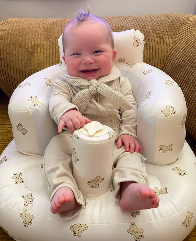 Baby in a support seat wearing a knitted outfit, with a beaming smile