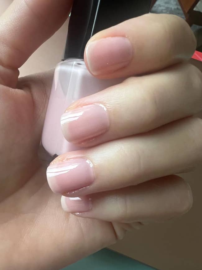 A hand holding a bottle of light pink nail polish, nails painted to match, showcasing a glossy manicure. Perfect for a chic, understated look