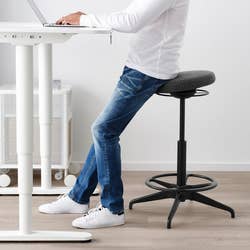 Person in jeans and white sweater sitting on a modern ergonomic stool at a standing desk, showcasing home office furniture