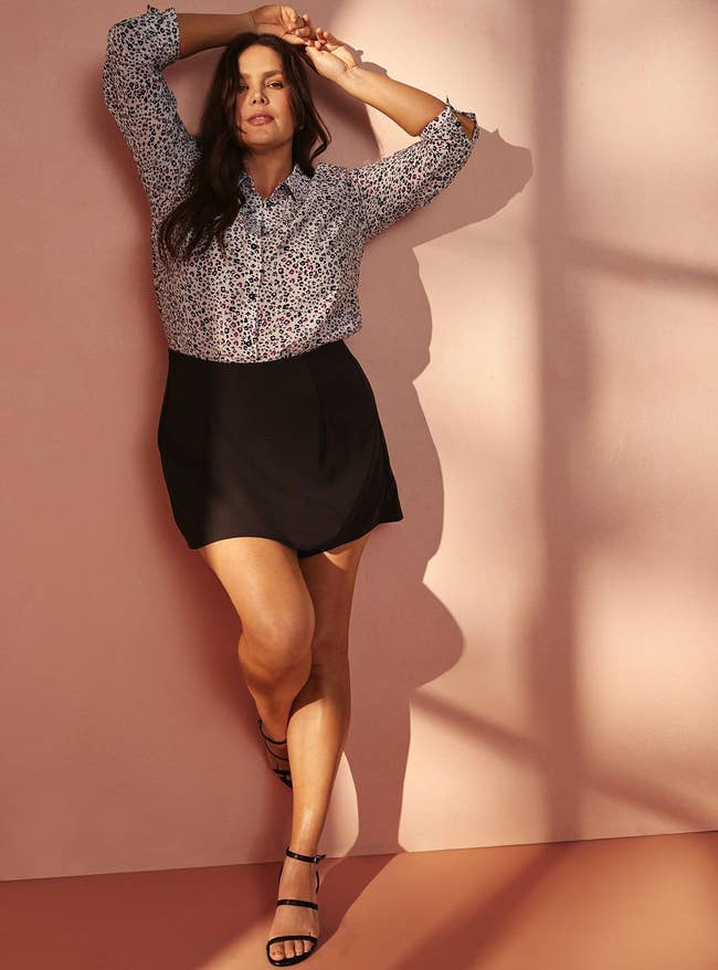 Person poses against a wall, wearing a patterned blouse, achromatic  skirt, and strappy sandals, suitable for a stylish and casual look