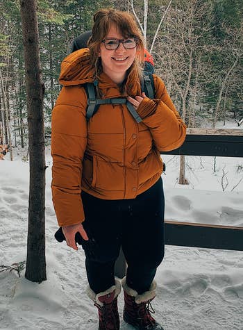 Person outdoors successful  a snowy setting, wearing a lukewarm  padded jacket, glasses, and carrying a backpack. They are smiling and dressed for wintertime  hiking