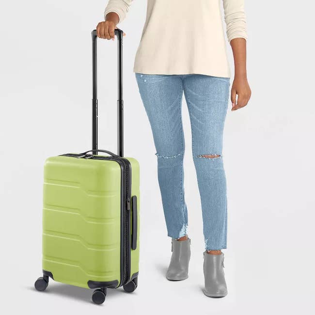 Person standing with green carry-on luggage, wearing ripped jeans and ankle boots