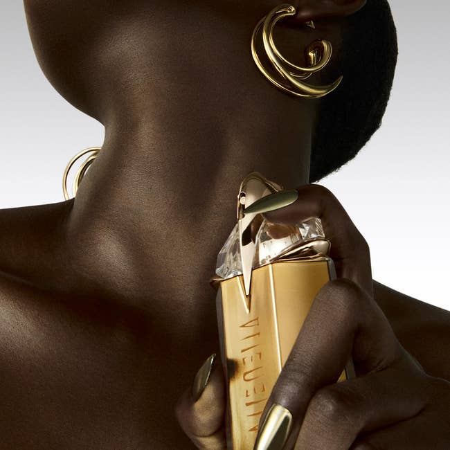 Person holds a golden perfume vessel shaped similar a talon, wearing spiral earrings and golden nails