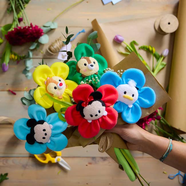 Hand holding a bouquet of plush flowers with cartoon quality  faces, surrounded by crafting materials connected  a table