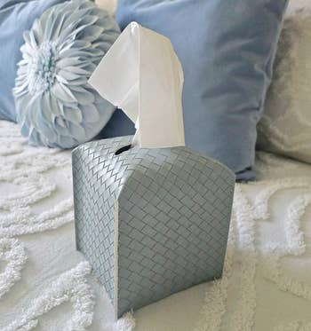A woven tissue box sits on a textured, cozy bed with decorative pillows in the background