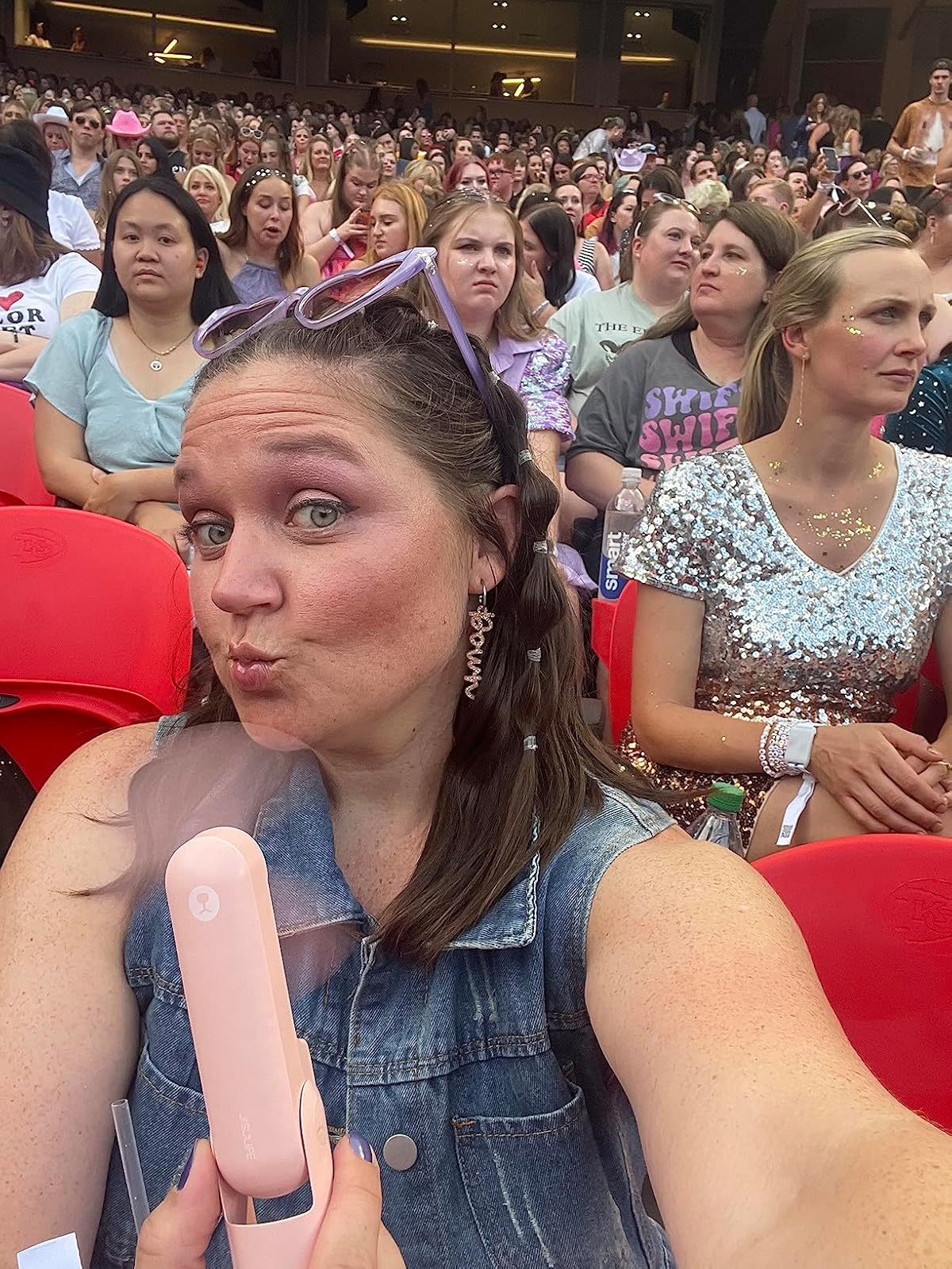 Person wearing a denim apical  poses for a selfie with a handheld fan, seated successful  a crowded stadium with attendees successful  casual and glittery attire