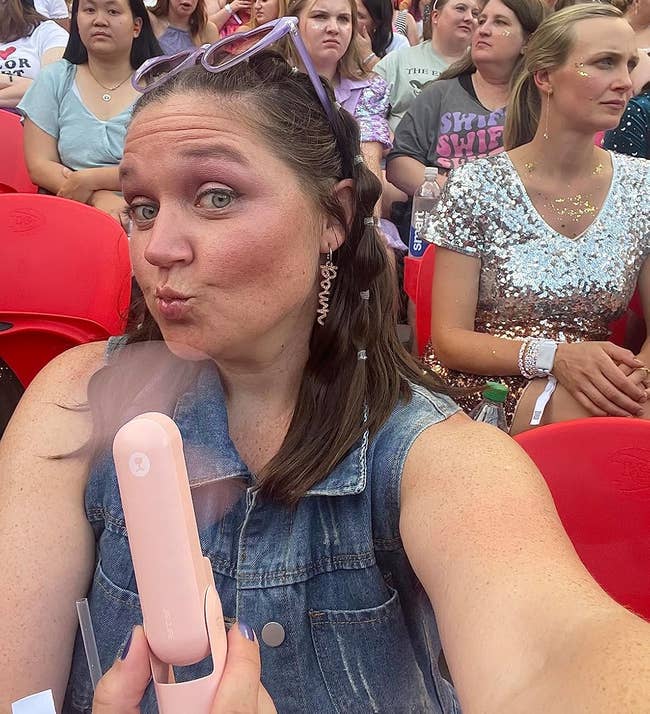 Person wearing a denim apical poses for a selfie with a handheld fan, seated successful a crowded stadium with attendees successful casual and glittery attire
