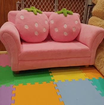 Child's pink sofa designed to resemble a cartoonish strawberry, with two fruit-themed cushions, placed on multicolored interlocking foam mats