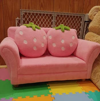 Child's pink sofa designed to resemble a cartoonish strawberry, with two fruit-themed cushions, placed on multicolored interlocking foam mats