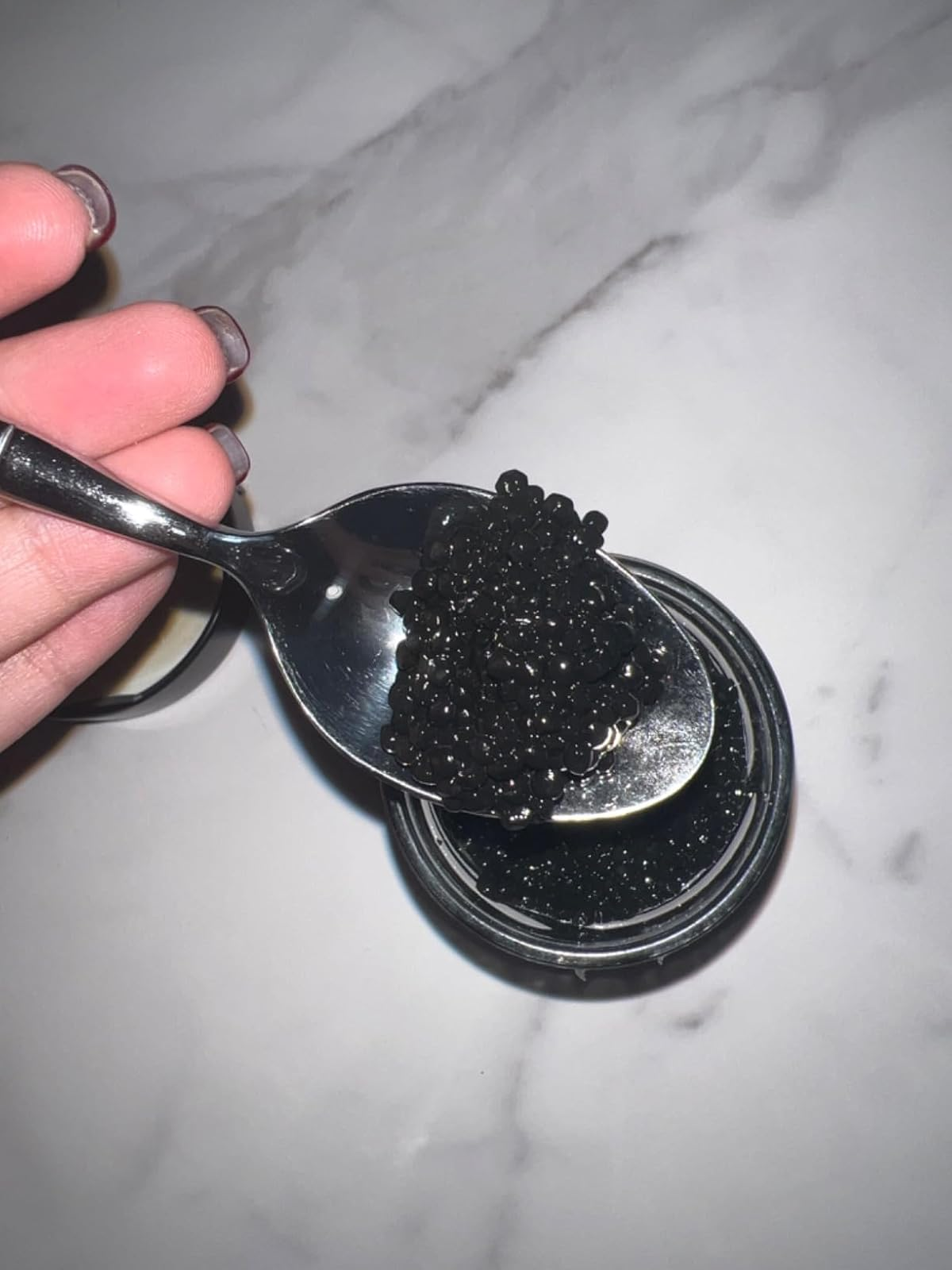 A hand holding a spoon of black caviar over an open jar 