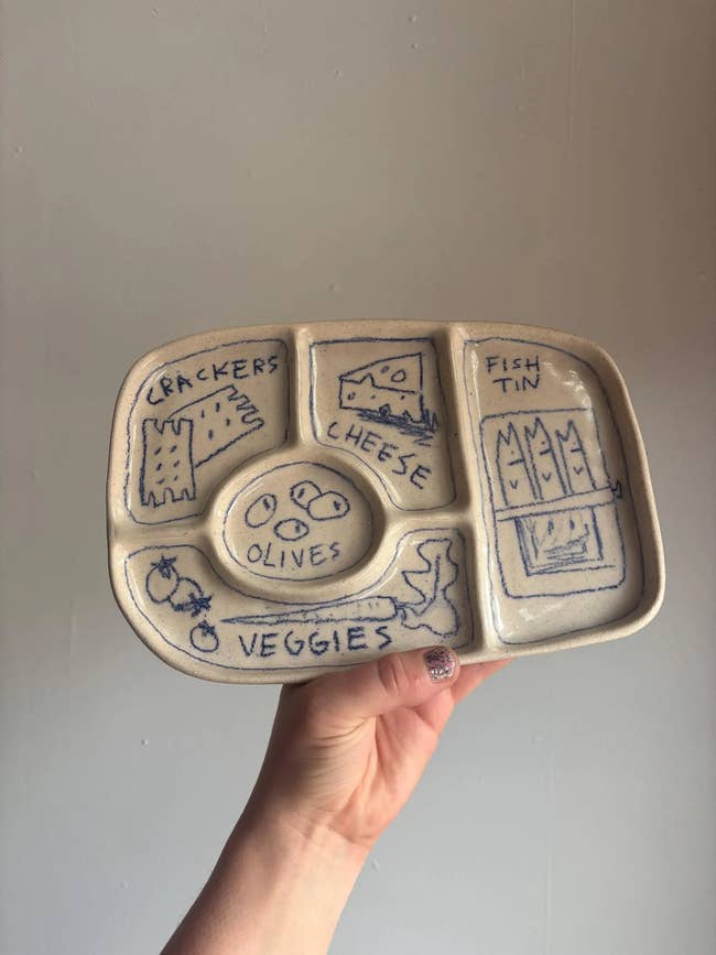 Hand holding a ceramic serving crockery with labeled sections for crackers, cheese, fish, olives, and veggies, perfect for snacks oregon appetizers
