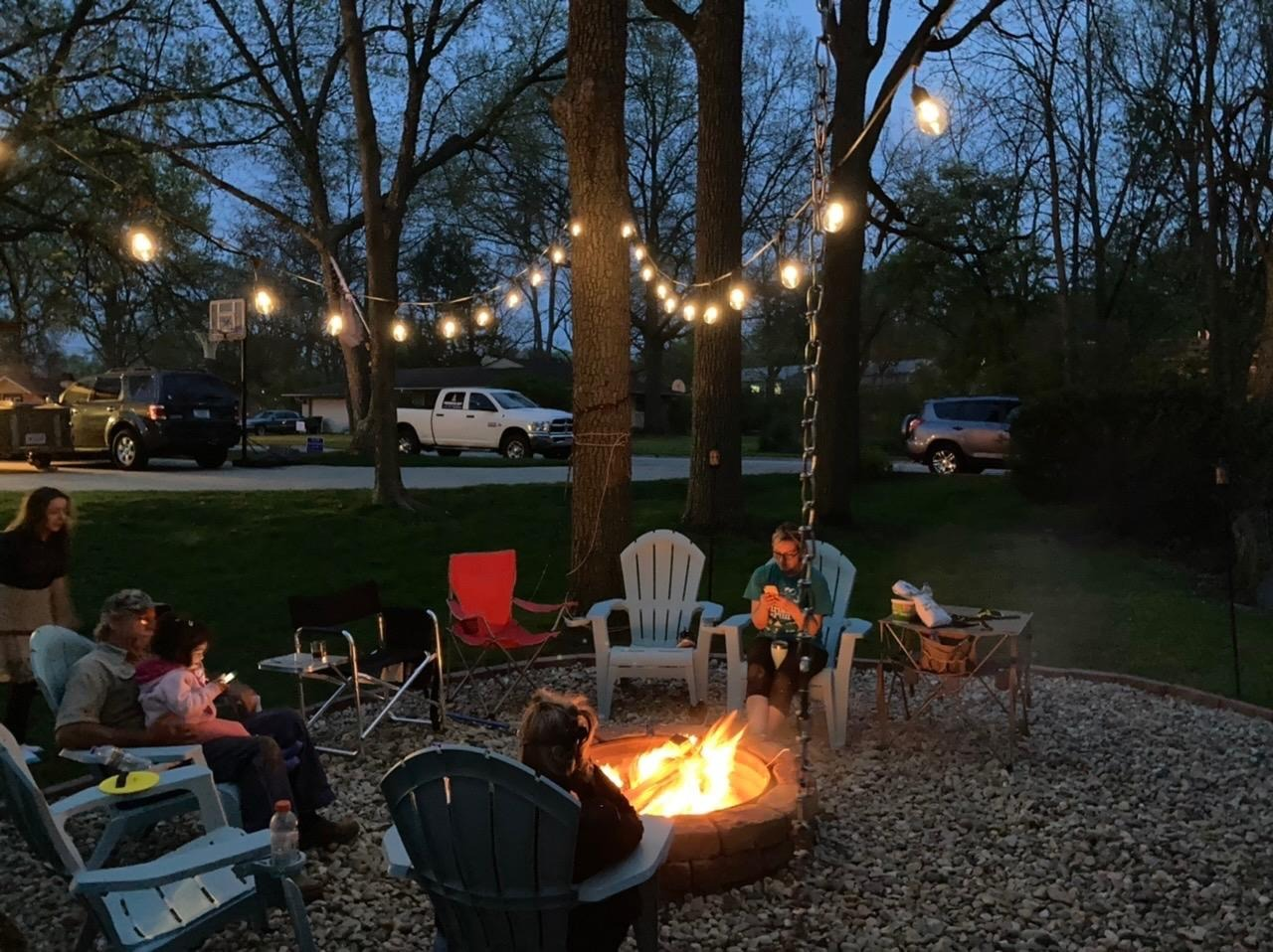 Family seated astir   a occurrence  pit successful  a backyard astatine  dusk, nether  drawstring  lights, with trees and parked vehicles successful  the background