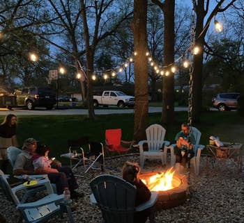 Family seated astir a occurrence pit successful a backyard astatine dusk, nether drawstring lights, with trees and parked vehicles successful the background
