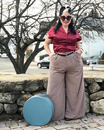 reviewer standing by stone wall, wearing 40s inspired clothes, including plaid wide palazzo pants