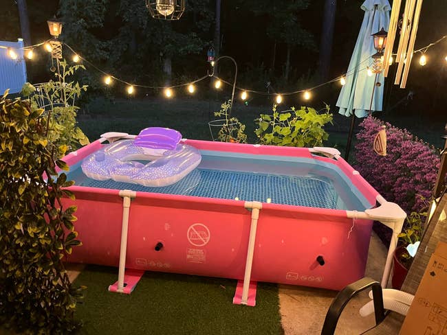 An inflatable excavation  with a purple interval  is connected  a patio with drawstring  lights, surrounded by plants and a plot  umbrella