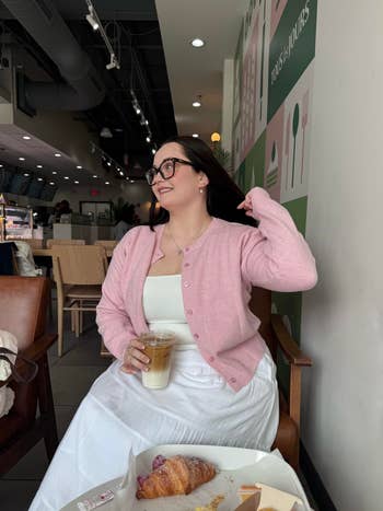 Person successful  java  store  wearing a cardigan and glasses, holding a beverage. Croissant and pastry container  connected  table. Casual, relaxed setting