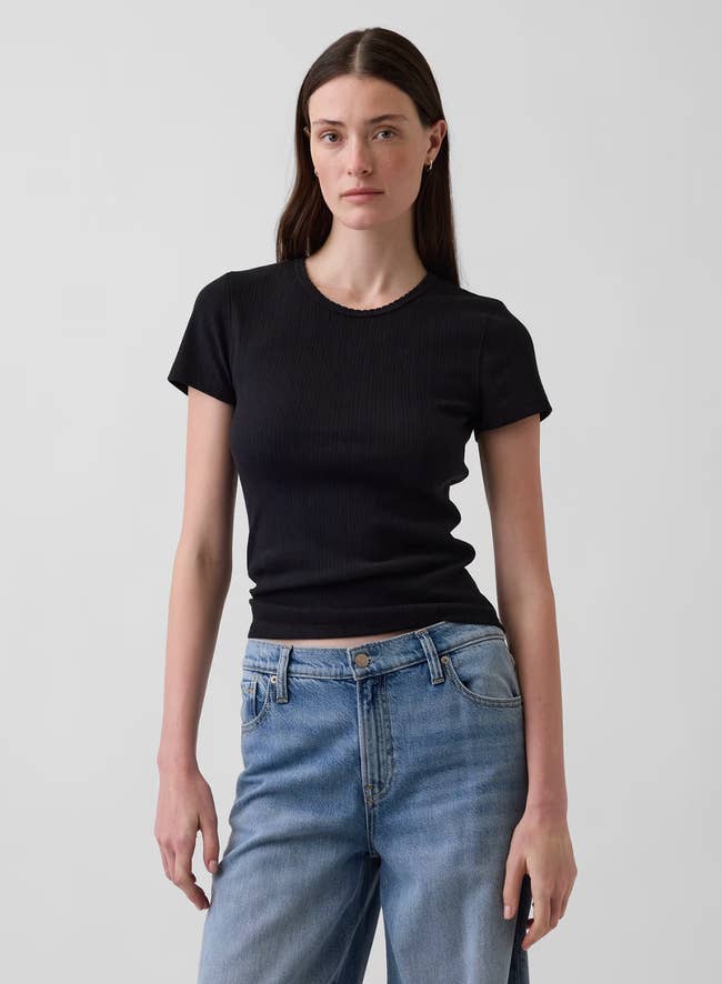 A idiosyncratic   wears a fitted achromatic  T-shirt and bluish  jeans, lasting  confidently against a plain background