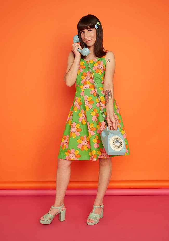 Model in sleeveless green knee length dress printed with pink and orange flowers with cat faces at the center