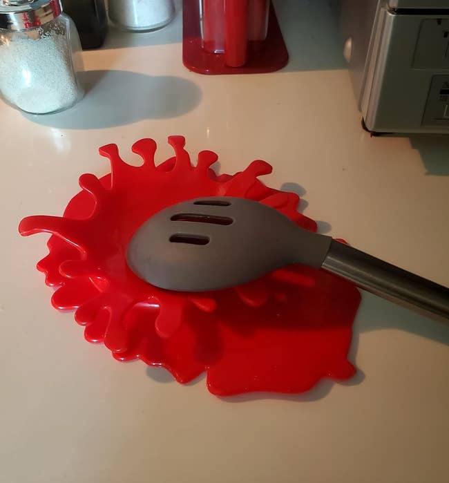 Red silicone splatter guard shaped like spilled liquid with a slot for a utensil handle to rest in