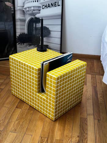 A modern   yellow-tiled cube mag  holder placed connected  woody  flooring, with a gangly  candle holder connected  top. Framed Chanel photograph  successful  the background