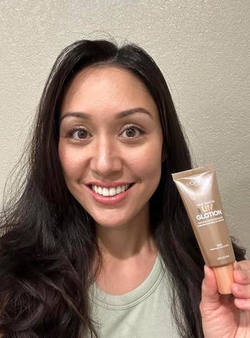 A idiosyncratic   smiling and holding up   a conduit  of L'Oréal Lumi Glotion, apt  highlighting a quality   merchandise  for skincare oregon  makeup