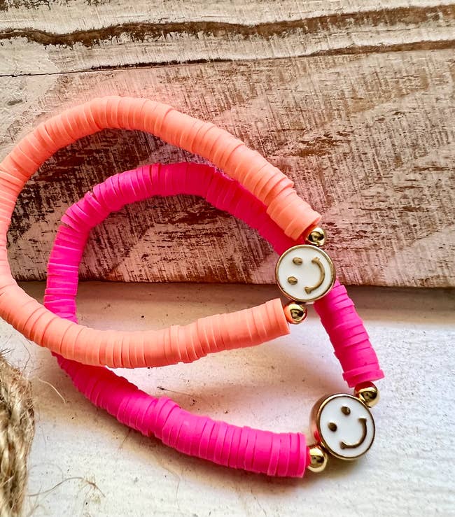 Orange beaded bracelet with two gold beads and a gold and white smiley face charm next to product in pink on a cement table