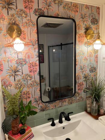 Bathroom with tropical-themed wallpaper, a black-framed mirror, golden  partition  sconces, achromatic  faucet fixtures, greenish  tile backsplash, and assorted  decorative items