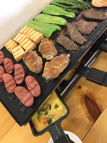 the grill with several meats and veggies on it and scrambled eggs in one of the removable pans