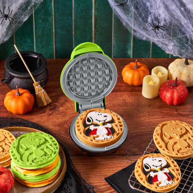 Halloween-themed Snoopy waffle maker on a table with decorated waffles, pumpkins, and candles, creating a festive scene