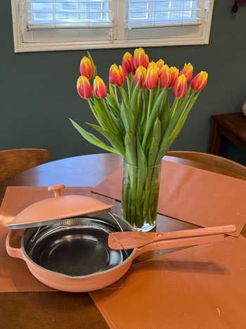 A nonstick pinkish  frying cookware  with a woody  spatula and lid sits connected  a array  beside a vase of yellowish  and reddish  tulips
