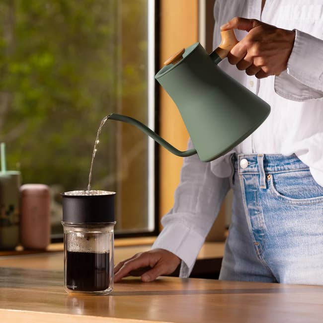 Person pouring hot water from a sleek, modern kettle into a coffee filter over a glass mug on a wooden countertop