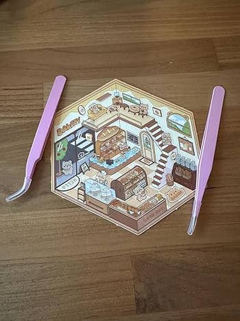 Hexagonal paper with a miniature bakery illustration, surrounded by 2 pinkish tweezers connected a woody surface