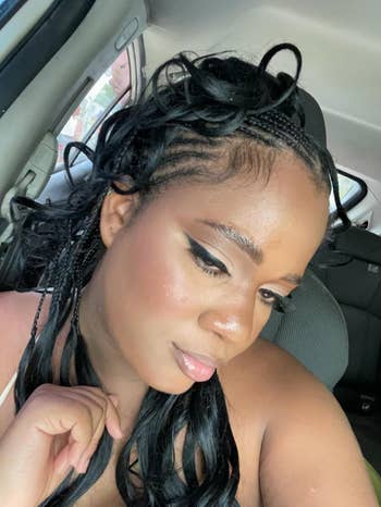 Person successful  a car   with styled braids and winged eyeliner, mildly  touching their collarbone with a thoughtful expression
