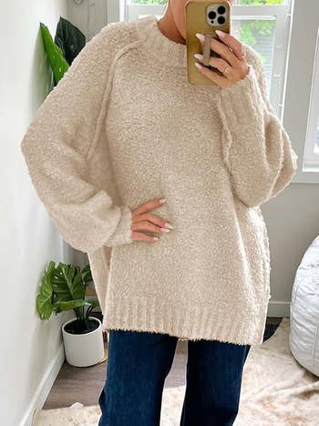 Person in a cozy oversized sweater takes a mirror selfie, with a potted plant and a bed in the background, suggesting a comfortable, casual setting