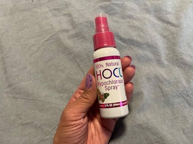 Person holding a vessel  of HOCl Hypochlorous Spray, labeled 100% natural, perfect  for skincare oregon  sanitizing, connected  a cloth  background