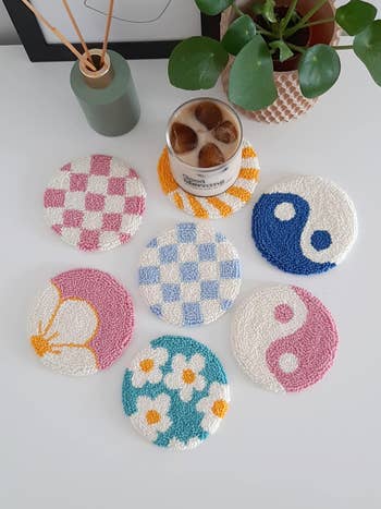 seven punch needle coasters in various designs including floral, yin and yang, and checkered patterns