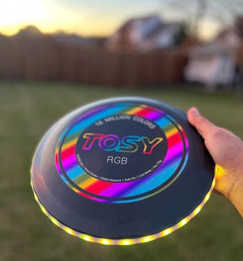 reviewer holding the black multi-color frisbee outside with the lights bright and visible in broad daylight