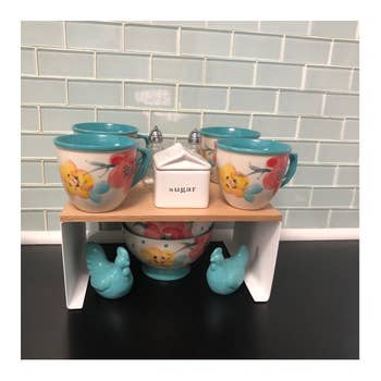 Set of floral mugs and sugar jar on a wooden shelf with a ceramic rooster and hen below, framed by a tile backsplash