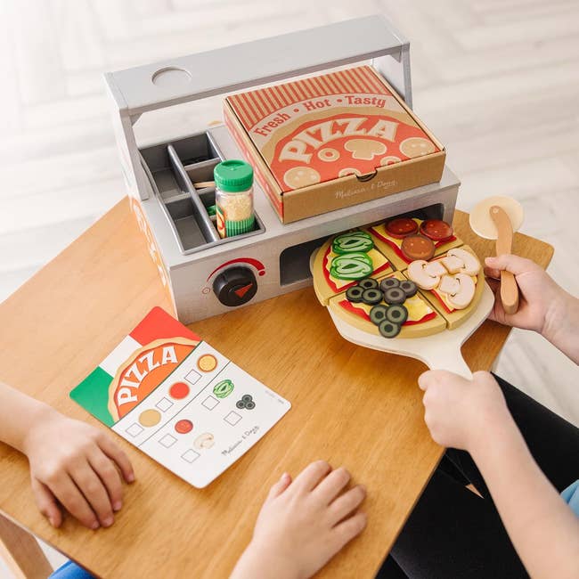 Children playing with a woody pizza artifact set, assembling slices and toppings. Toy oven and pizza container included