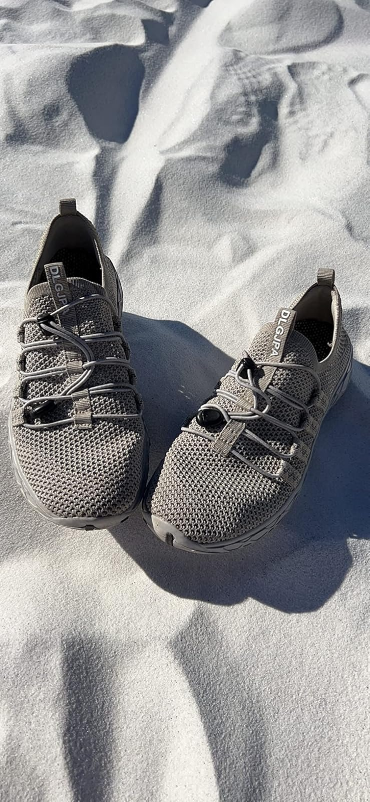Pair of grey  all-terrain shoes with knitted mesh uppers and elastic laces, positioned connected  achromatic  sand